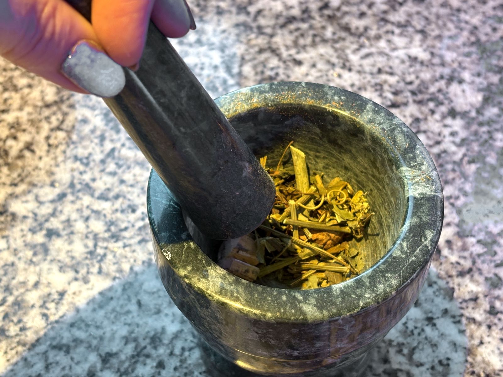 A hand grinding dried herbs with a pestle in a green marble mortar on a granite counter